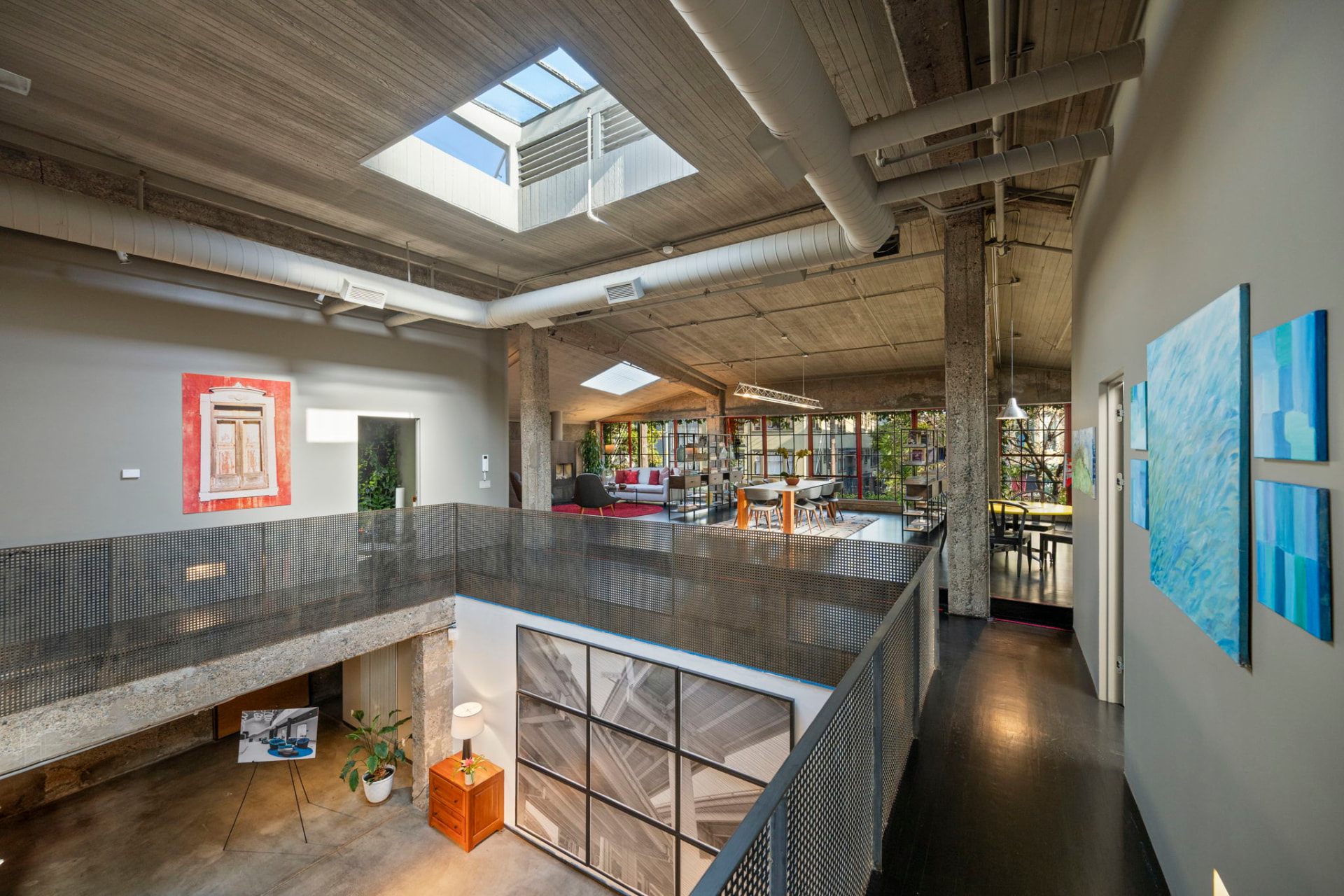 View from the kitchen mezzanine across the open loft toward tall red-framed factory windows and a dining area